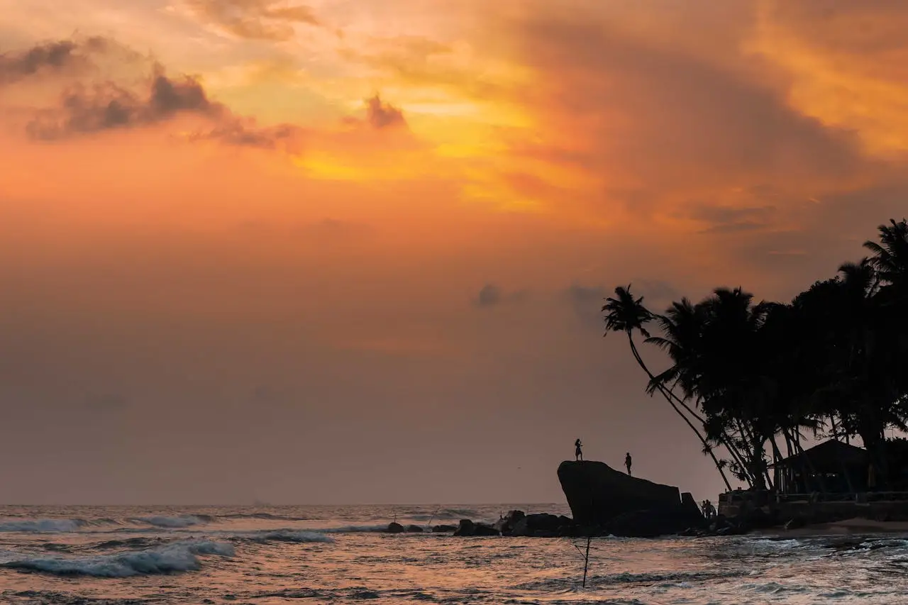 Dramatic sunset silhouette of two figures on a rocky shore at Unawatuna Beach, Sri Lanka.