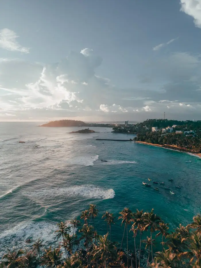 A stunning aerial view of Sri Lanka's coastline with palm trees, clear ocean waves, and a vibrant sunset sky.